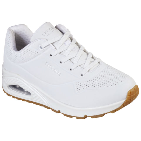 A white sneaker with a perforated design and a thick sole featuring a visible air cushioning unit is displayed in a neutral setting highlighting its clean aesthetic and sporty style.