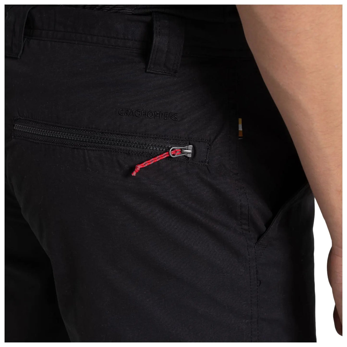 A pair of black hiking pants is displayed with a side view highlighting a zipper pocket that has a red pull tab situated just above the waistband and features a brand logo embossed on the fabric.