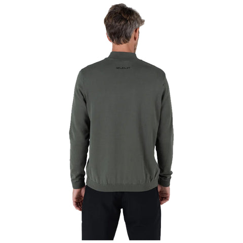 A man stands with his back to the viewer wearing a dark green sweater and black pants in a neutral space highlighting the clothing design and fit.