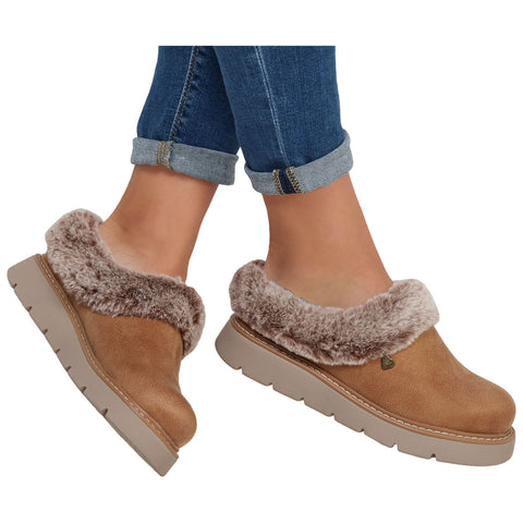 Brown slip-on shoes with a fluffy lining are being worn by a person whose jeans are rolled at the cuffs while standing on a neutral background.