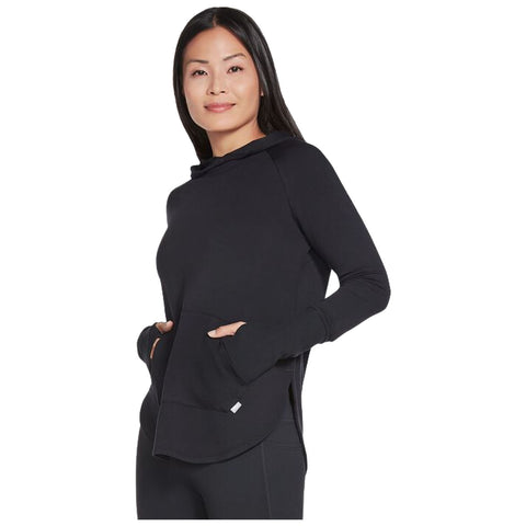 A woman stands wearing a black hoodie with a pocket smiling confidently her hands are partially visible in the pocket against a plain white background