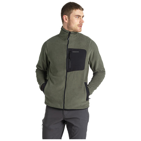 A man stands wearing a green fleece jacket with a black chest pocket and gray pants while looking to his left in a neutral background setting.