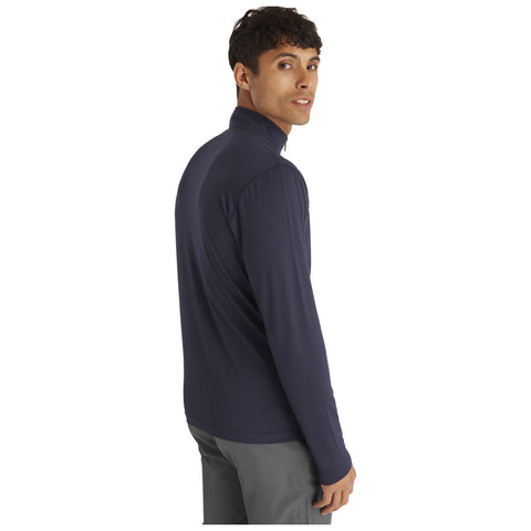 A man wearing a navy long-sleeve shirt is standing with his body turned slightly away from the viewer while looking back over his shoulder in a neutral background.