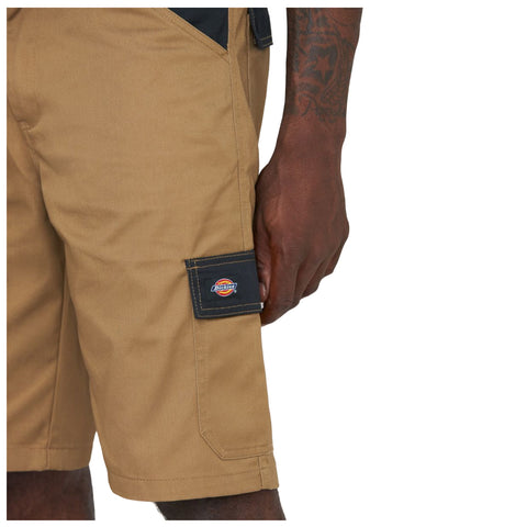 Tan work shorts sit above the knee with a black patch on the side pocket displaying a logo while a hand rests casually on the thigh area in a neutral setting.