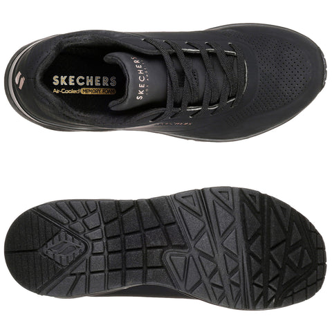 Black sneakers are positioned side by side with one foot slightly raised showcasing their design while a person wearing olive green pants stands in a neutral setting highlighting the footwear.