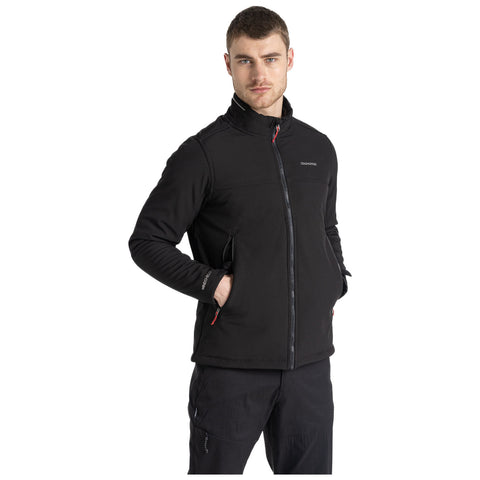 A man stands confidently wearing a black zip-up jacket with a high collar and dark pants, placing his hands in the jacket pockets against a plain white background.