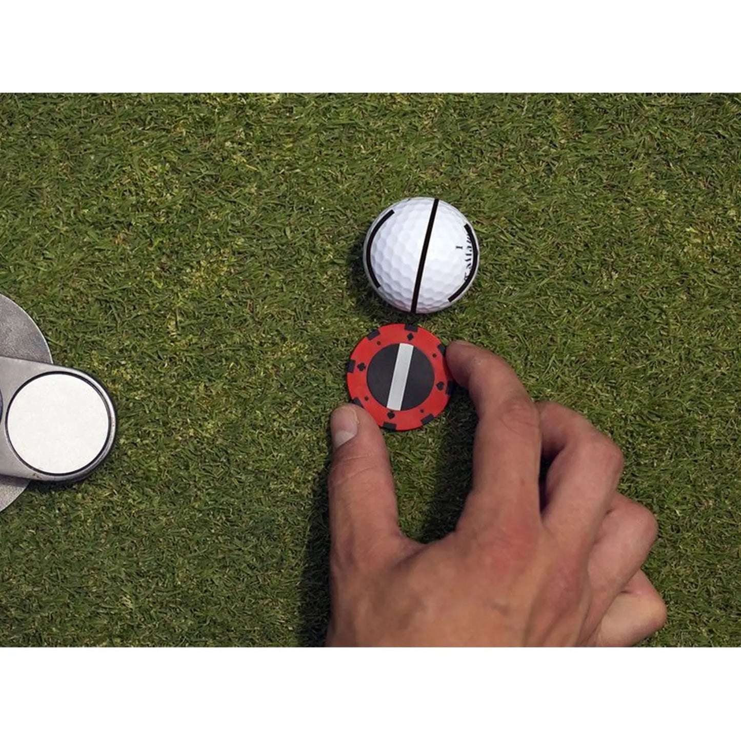 A hand is placing a red marker with a black line on the green grass next to a white golf ball with black stripes while a silver putting device is nearby