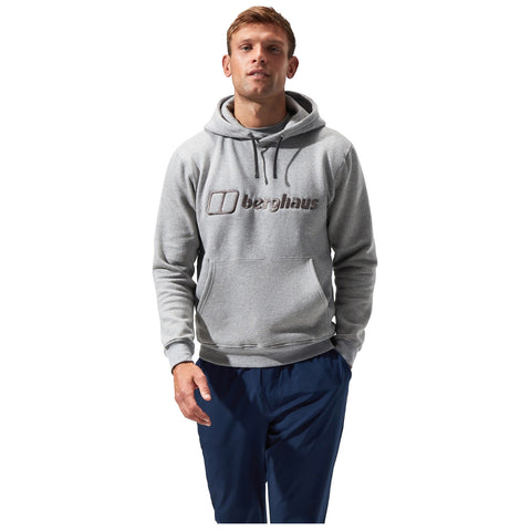 A man stands confidently wearing a grey hoodie with a logo on the chest and navy pants in a neutral background, suggesting a casual, outdoor-ready appearance.