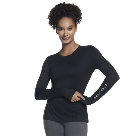 A woman with curly hair is posing confidently while wearing a fitted long-sleeve black top and grey pants with the word SKECHERS printed on her sleeve in white.