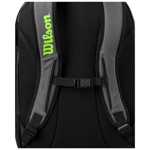 A gray backpack features padded mesh shoulder straps with a green Wilson logo prominently displayed the straps are secured with an adjustable buckle against a black mesh background.