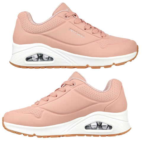 Pink athletic shoes with a cushioned sole are positioned to showcase different angles. They feature a smooth finish, perforated sections, and prominent branding on the side.