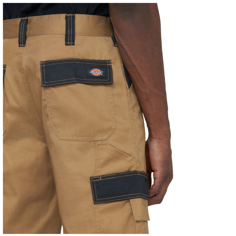 Khaki cargo pants are shown with black accents and pockets The person wearing them is holding the waistband with one hand while their other arm is relaxed by their side