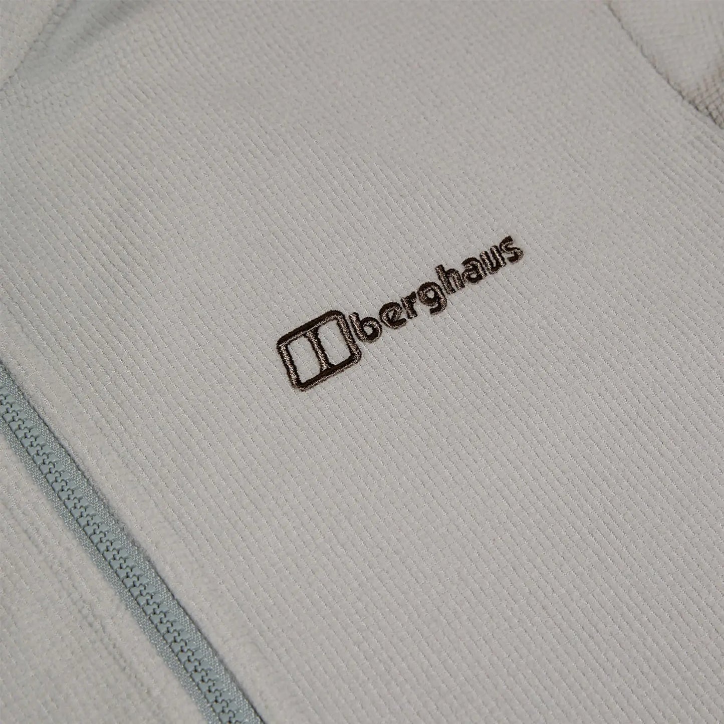 A light-colored fleece garment lies flat displaying a textured fabric with a zipper on one side featuring the embroidered brand name Berghaus in brown near the upper left.