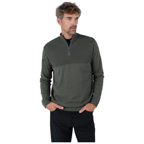 A man wearing a green zip-up sweater stands with his hands in his pockets looking relaxed and contemplative against a plain white background.