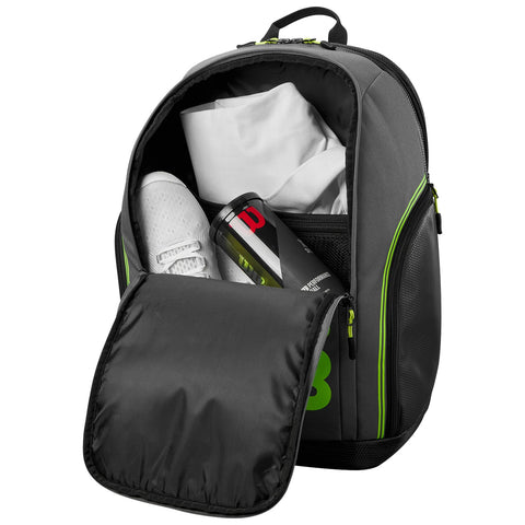 A gray backpack with green accents is partially open revealing a pair of white sports shoes a black can and a folded white shirt inside in a casual indoor setting