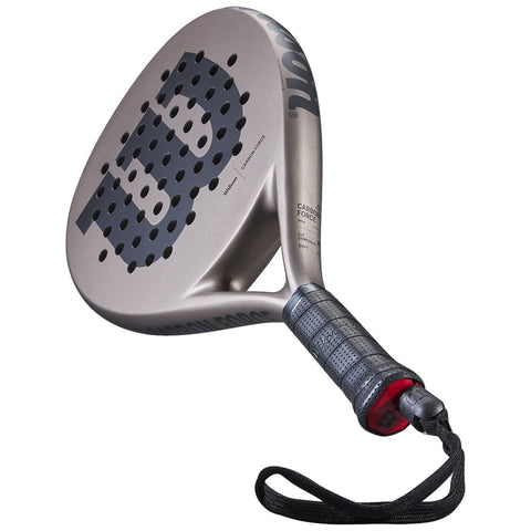 A padel racket stands upright showing a perforated hitting surface and a textured grip. The context suggests it is designed for outdoor sports play, likely on a court.