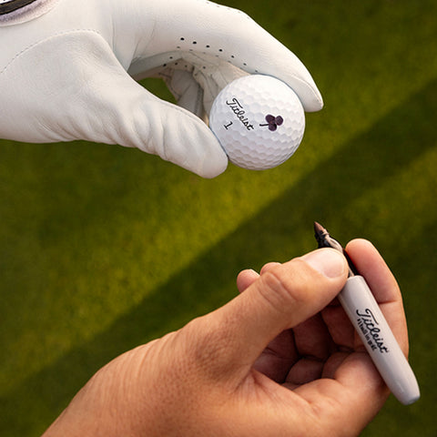 A gloved hand holds a Titleist golf ball while another hand with a marker prepares to write on it in a green outdoor setting.