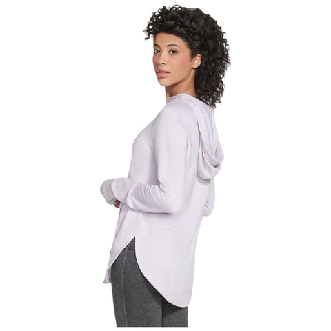 A woman wearing a light purple long-sleeve hoodie stands with her back turned, looking over her shoulder in a neutral background. She is styled with curly hair and wearing gray pants.
