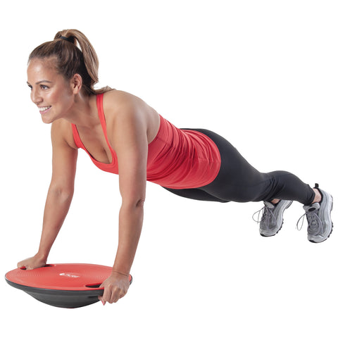 A woman in a red tank top performs a plank exercise while gripping a round red balance disc in a neutral environment highlighting physical fitness and strength training.
