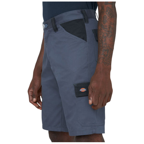 Gray cargo shorts are being worn by a person who stands with one hand near the pocket displaying a logo on the side in a neutral background setting.