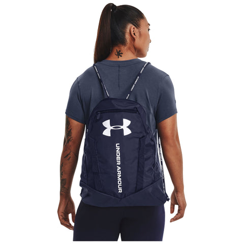 A person wears a dark blue backpack with a prominent white logo and text, standing in a casual attire, showcasing a tattoo on their neck in a neutral environment.