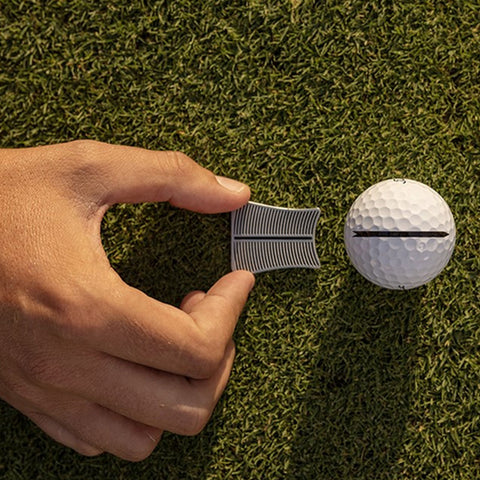 A hand holds a gray golf tee while positioning it next to a white golf ball on green grass preparing for a shot in a golfing context.