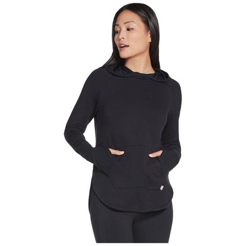 A black hoodie is being worn by a woman with long hair She is standing with her hands in the front pockets looking slightly to her left in a neutral setting