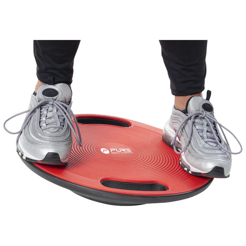 A person stands on a red balance board with curved edges, their feet positioned securely while balancing. The environment suggests an exercise or fitness setting.