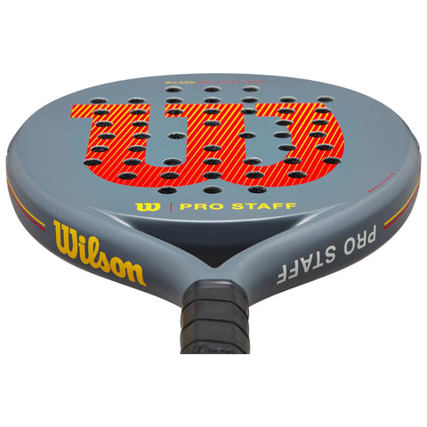 A grey paddle racket rests on its handle displaying a prominent red and yellow Wilson logo on its face surrounded by numerous holes designed for performance and grip.