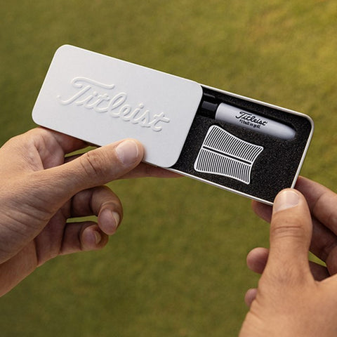A hand holds a white box with "Titleist" embossed on it containing a marker and a comb sitting on black foam, set against a green grass background.