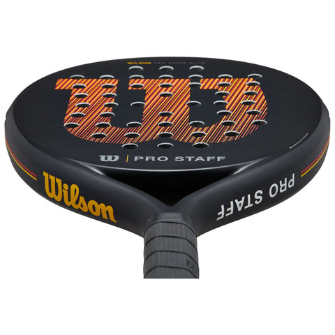 A black paddle with a circular head displays the "Wilson" logo in bold orange and yellow stripes. It features several perforations and a textured handle for grip.