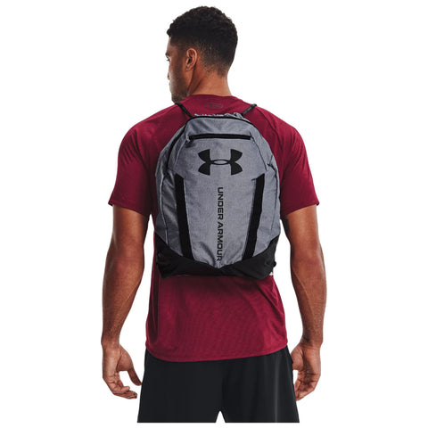 A man wearing a red shirt stands with a black and gray Under Armour backpack secured on his back in a neutral setting likely indoors or outdoors.
