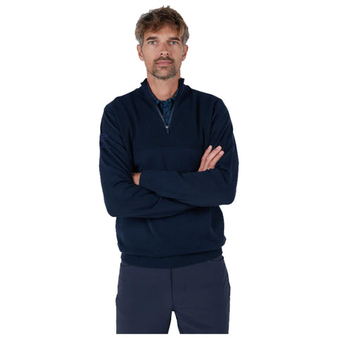 A man with short hair wears a dark navy quarter-zip sweater and stands with arms crossed. He presents a composed expression against a plain background.