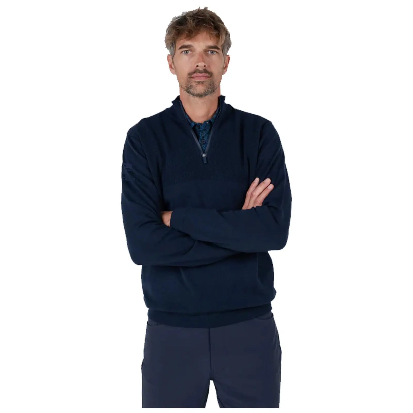 A man with short hair wears a dark navy quarter-zip sweater and stands with arms crossed. He presents a composed expression against a plain background.