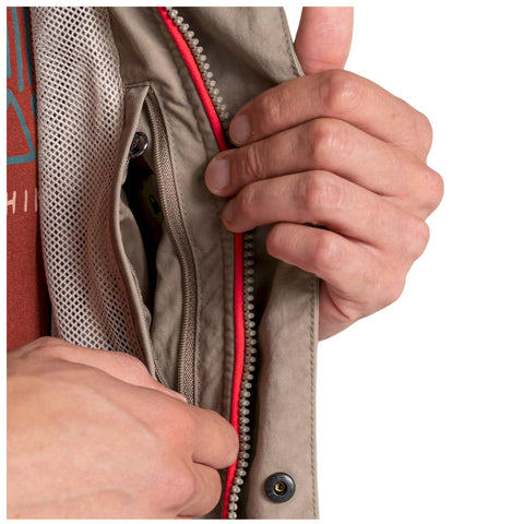 A person is reaching into a zipped pocket of a beige jacket using one hand while the other hand holds the edge of the pocket. The jacket features a red zipper and breathable mesh lining.