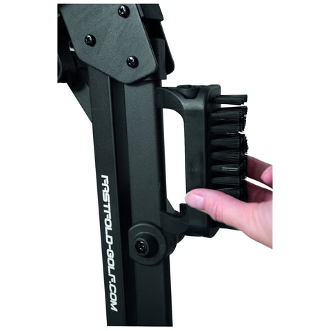 A hand is gripping a small brush attached to a black golf accessory which is designed for cleaning clubs, located in a workshop or outdoor setting intended for golfers.