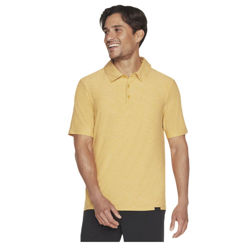 A smiling man wears a short-sleeved yellow polo shirt and stands with one hand raised slightly in a casual pose against a plain background.
