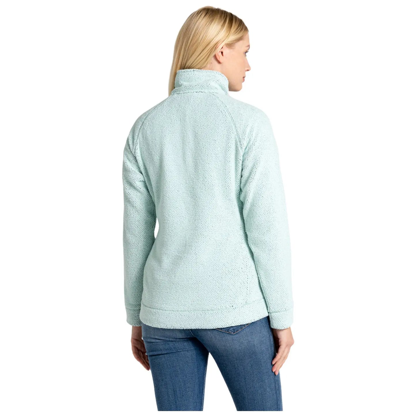 A woman is wearing a light blue fleece jacket with a high collar while standing in a relaxed pose. She has long blonde hair and is facing away from the viewer.
