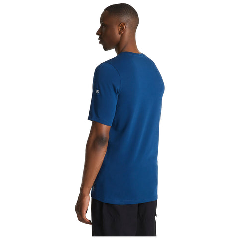 A man stands facing slightly away wearing a blue short-sleeved shirt and dark pants in a neutral background. The shirt features a small logo on the sleeve.