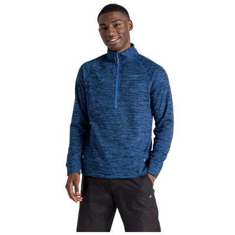 A man stands facing forward wearing a navy blue zip-up fleece top with a small logo on the chest and light-colored trousers in a plain white background