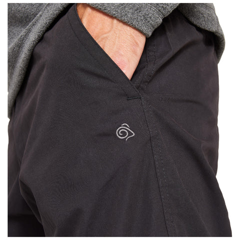A person wearing grey clothing has their hand in the pocket of black pants featuring a small spiral logo on the side in a casual indoor setting.