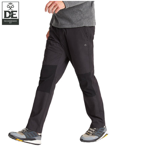 A person is walking while wearing dark outdoor pants with tonal patches and sturdy shoes in an active posture against a simple white background with a logo in the top left corner.