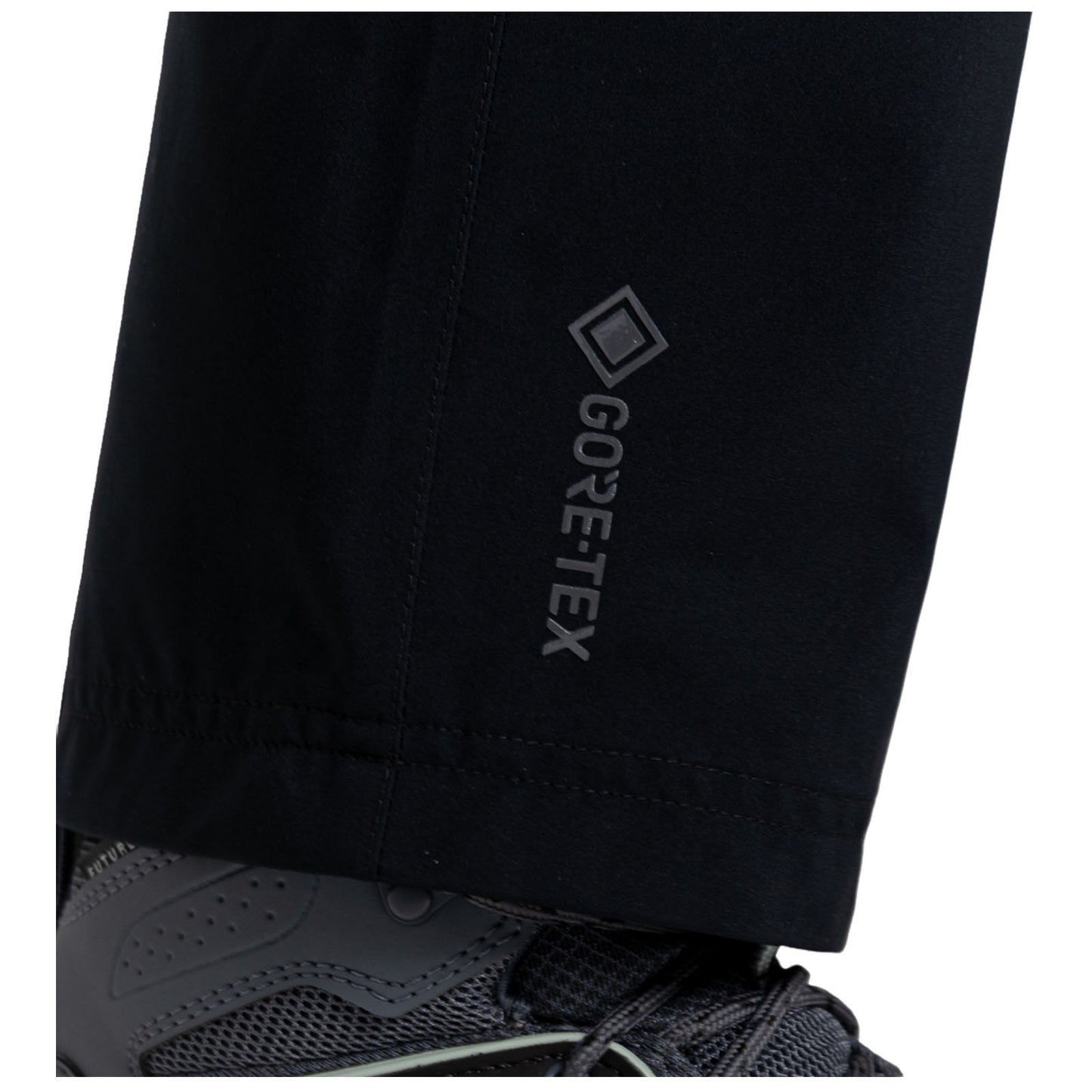 Black waterproof pants with a GORE-TEX logo are partially visible above a dark athletic shoe featuring a textured surface and reinforced toe, set against a plain background.