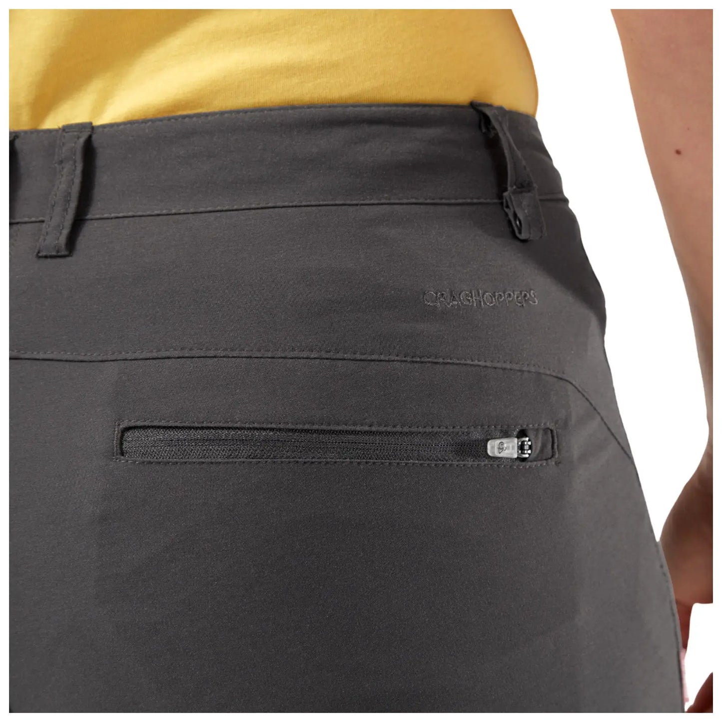 A pair of dark gray shorts is displayed with a visible side zippered pocket and a small logo stitched on the waistband while a person wearing a yellow shirt stands behind them.