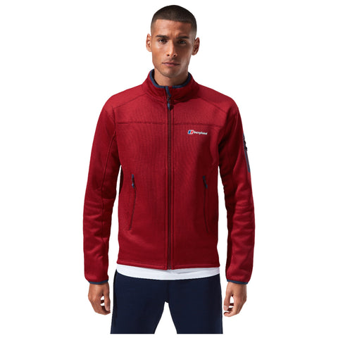 A man stands upright wearing a red zip-up jacket with a brand logo visible on the left chest area and navy blue accents presenting a casual clothing style in a neutral background.