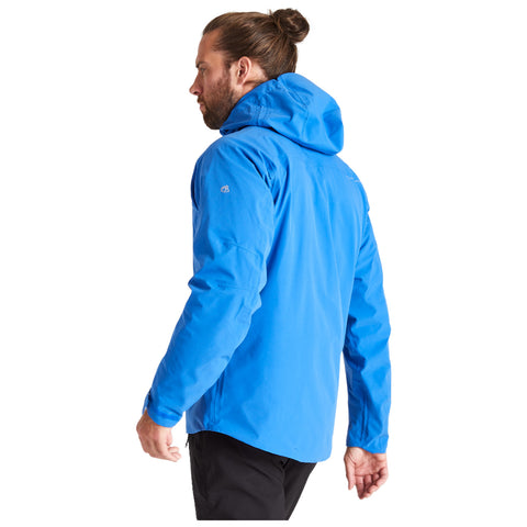 A man is turning slightly to his left while wearing a bright blue jacket with a hood in a neutral background without distractions. His hair is tied back in a bun.