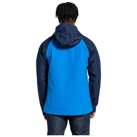 A person is standing with their back facing the viewer wearing a two-toned blue and navy jacket in a plain white background. The individual has curly hair and dark jeans.