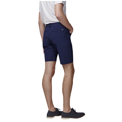 A person stands with their back facing the viewer wearing navy shorts and a white shirt paired with dark shoes in a neutral setting with no distinct surroundings visible.