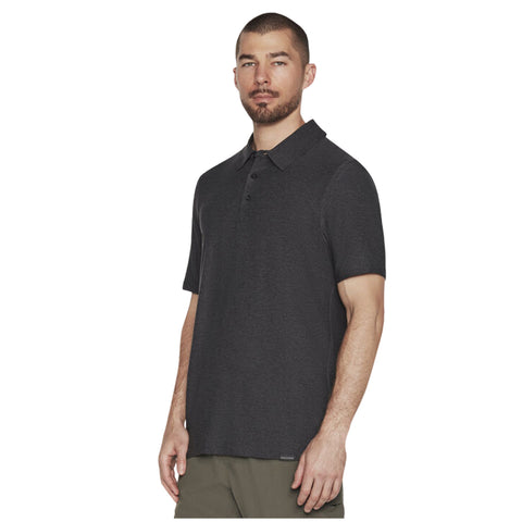 A man stands confidently wearing a dark gray short-sleeved polo shirt paired with olive green pants in a neutral background, posing with a serious expression.