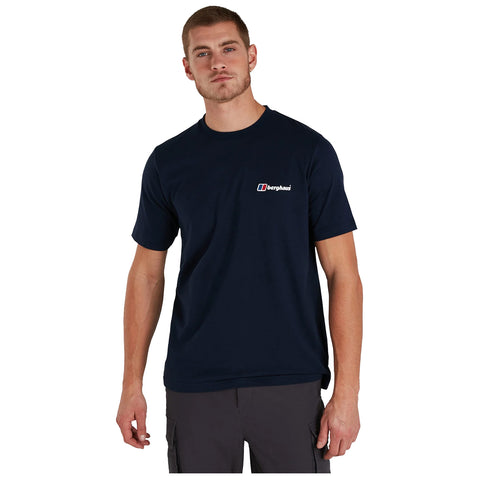 A man stands confidently wearing a navy blue t-shirt with a small logo on the chest and gray cargo shorts in a neutral background.